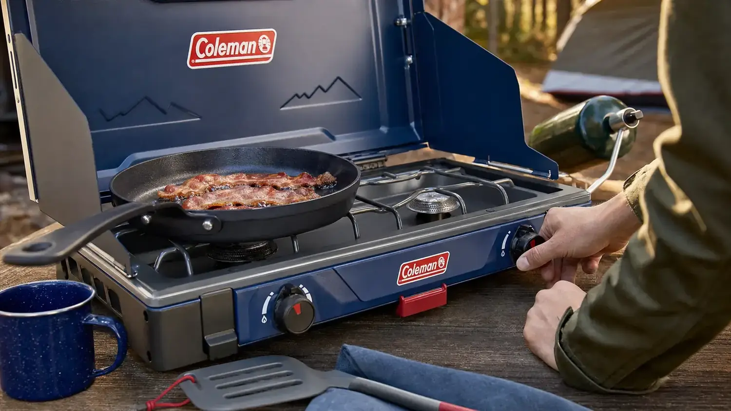 Hand adjusting the burner knob on a propane camp stove while a pan of bacon sizzles on the grate