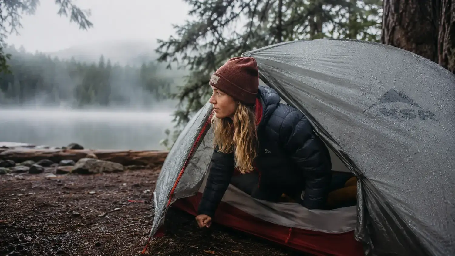 Camper opening the Coleman Sundome Dark Room tent door at a lakeside campsite on a foggy morning, rain fly still beaded with water from overnight rain