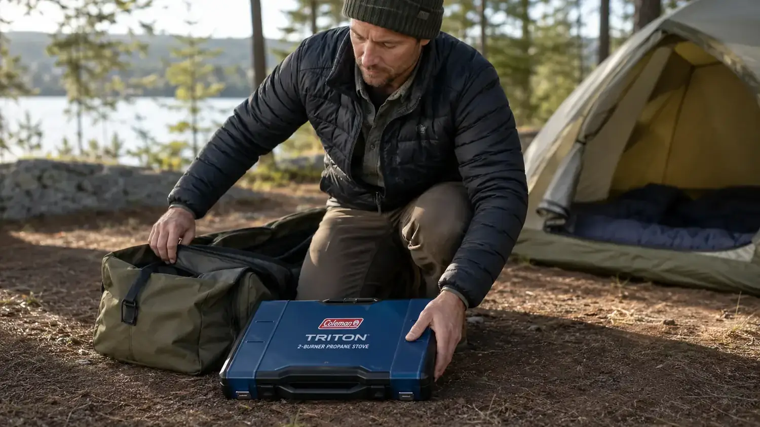 Two-burner propane camp stove packed flat in its carry case on the ground next to a camp bag