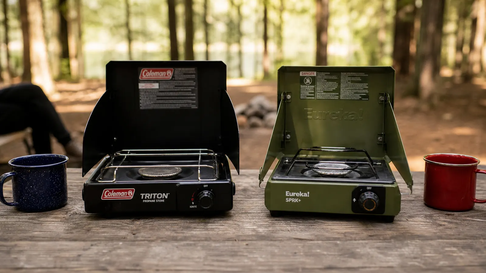 Two two-burner propane camp stoves side by side on a picnic table at a forested campsite