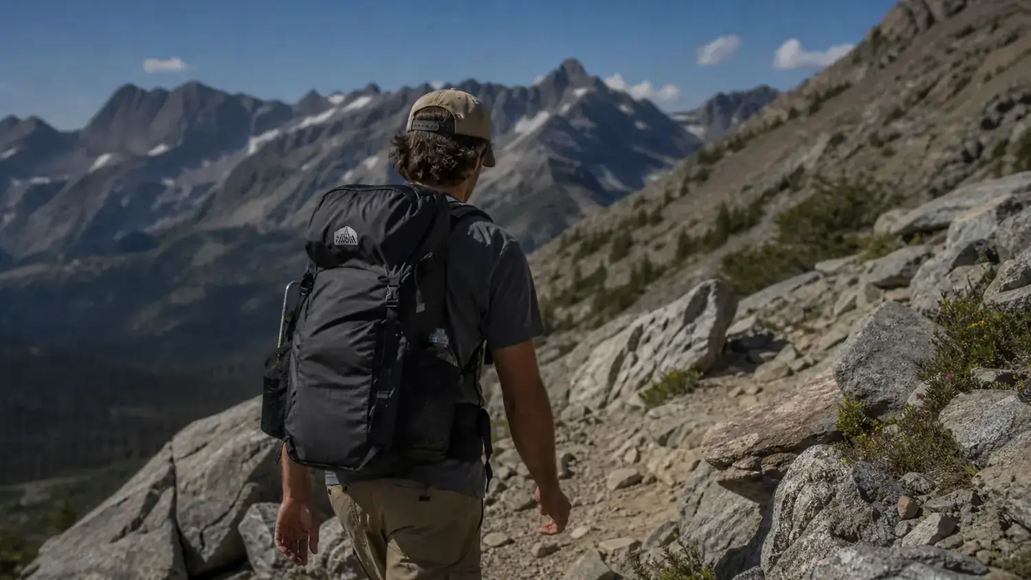 Hiker with a loaded backpack crossing a rocky trail through alpine meadow, pack sitting high and close to the body