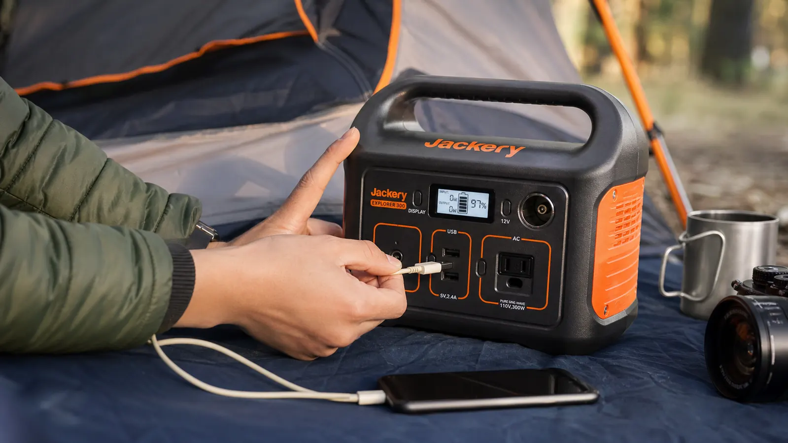 Hand plugging a phone charging cable into the USB port of a Jackery Explorer 300 resting on a camp tarp next to a tent